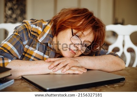 Tired mature lady in glasses lying down on laptop at desk in home room. Advanced pensioner practicing computer skills and learning modern technology. Online communication and distant future concept