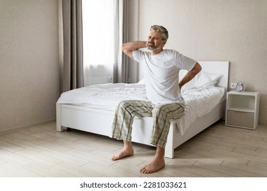 Tired mature man suffering from back pain after sleeping, waking up in morning, sitting on bed and touching his lower back and neck, experiencing body aches, wearing pajamas, copy space, full length - Powered by Shutterstock