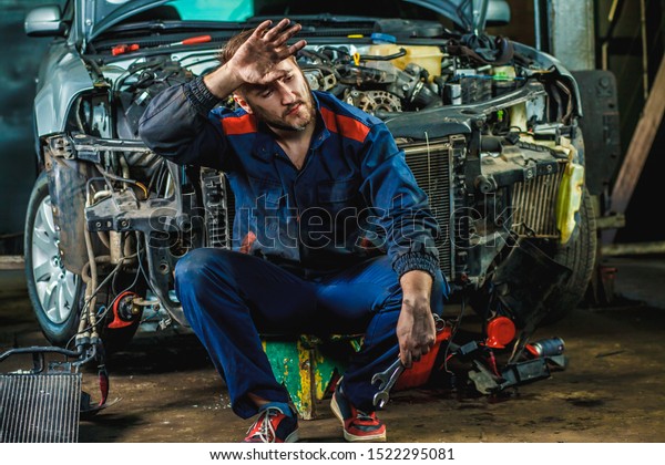 A tired\
mechanic in a blue protective suit is sitting near a disassembled\
car. Repair Service Concept. 