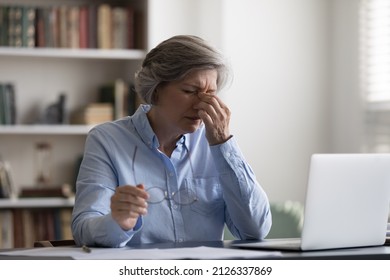 Tired middle aged woman taking off eyeglasses, suffering from painful feelings or having blurry eyesight, having astigmatism problem. Stressed due to long computer workday old lady relieving eyes pain