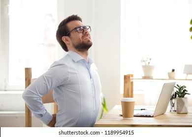 Tired Millennial Businessman Suffering From Backache Feeling Badly, Sitting At Office Desk Indoors. Have A Back Pain, Sitting At The Computer For A Long Time, Sedentary Work And Bad Posture Concept