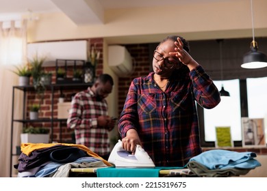 Tired Modern African American Wife Ironing Clothes While Relaxed Distracted Husband Uses Cellphone. Disappointed And Exhausted Wife Frustrated At Taking Care Of All Household Chores Alone Without Help