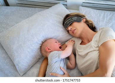 Tired mother with eye band trying to sleep while little toddler crying on bed. Angry baby boy crying and trying to wake mother. Stressed mother trying to take a nap with her child.