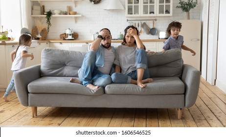Tired mother and father sitting on couch feels annoyed exhausted while noisy little daughter and son shouting run around sofa where parents resting. Too active hyperactive kids, need repose concept