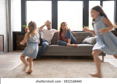 Tired Mother And Father Sitting On Couch Feels Annoyed Exhausted While Noisy Daughters Shouting Run Around Sofa