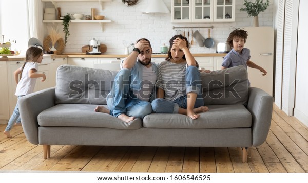 Tired mother and father sitting on couch feels\
annoyed exhausted while noisy little daughter and son shouting run\
around sofa where parents resting. Too active hyperactive kids,\
need repose concept