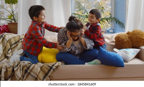 Tired Mother Feels Exhausted While Noisy Little Kids Annoying Her. Tired Young Woman Nanny Sitting On Couch With Two Naughty Boys Beating Her And Misbehaving