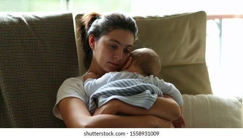 
Tired Mother Holding And Caring For Baby Infant On Couch. Exhausted Mom Falling Asleep, Candid And Authentic