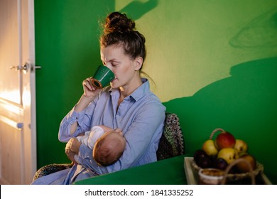 Tired Mother In Kitchen At The Table Puts Newborn To Sleep. Mom Rocks The Baby And Sings A Lullaby. Caring For Baby Hug Tenderness. Sleep Deprivation Insomnia Drink Coffee. Night Postpartum Depression