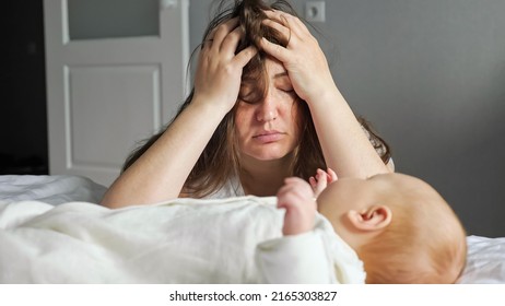 Tired mother looks after little newborn child trying to ignore headache. Little baby girl lies on bed with white quilt and looks at suffering mother - Powered by Shutterstock