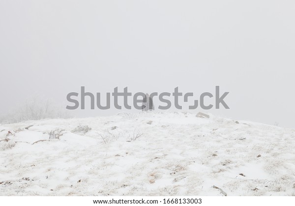 Tired mountain hiker with hood over\
head walking through the misty, snow covered\
highlands