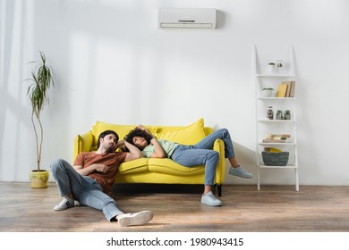 tired multiethnic couple sitting on yellow sofa and suffering from heat in summer