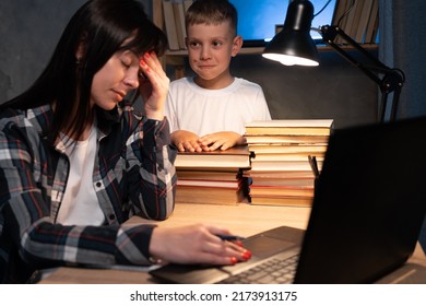 Tired Multitasking Mother Working From Home. Mom And Son Use Technology At The Table. Family Working On Laptop At Night Overtime