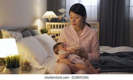 tired new mother sitting by lamp is dozing off while bottle feeding her newborn child at midnight in the bedchamber.