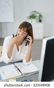 Tired Office Woman Sitting At Her Table, Talking To Someone On Mobile Phone And Showing Yawning Gesture.