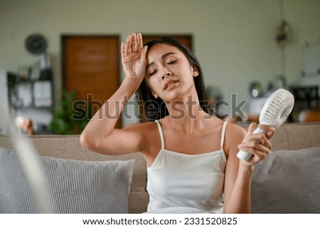 A tired and overheated Asian woman suffering from a heat attack uses an electric handy fan to cool herself down while resting on a couch in her living room on a hot summer day.