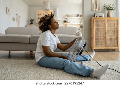 Tired overheated black woman cooling herself with electric fan at home, suffering from heat while sitting on floor in living room without conditioner, keeping cool indoors during summer heatwave