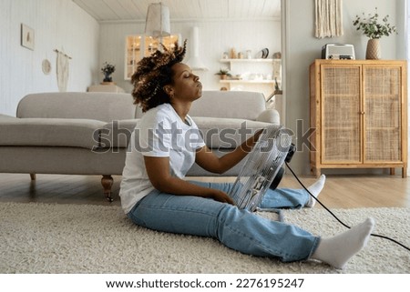 Tired overheated black woman cooling herself with electric fan at home, suffering from heat while sitting on floor in living room without conditioner, keeping cool indoors during summer heatwave