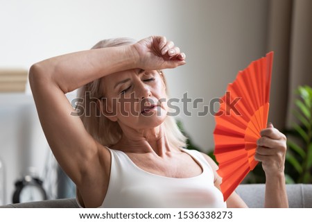Tired overheated middle aged lady wave fan suffer from menopause exhaustion complain on heat at home, stressed old woman sweat feel uncomfortable hot in summer weather problem without air conditioner