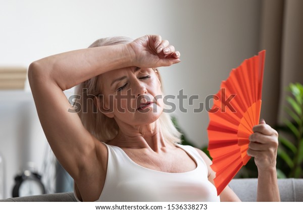Tired overheated middle aged lady wave fan\
suffer from menopause exhaustion complain on heat at home, stressed\
old woman sweat feel uncomfortable hot in summer weather problem\
without air conditioner