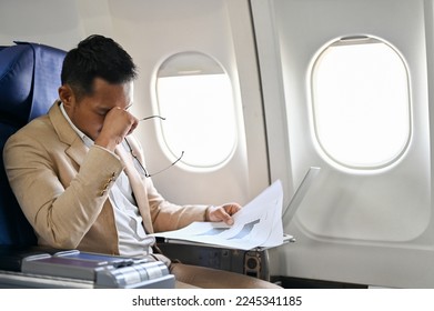 Tired, overworked and stressed Asian businessman reviewing financial report, working and managing his tasks during the flight. - Powered by Shutterstock