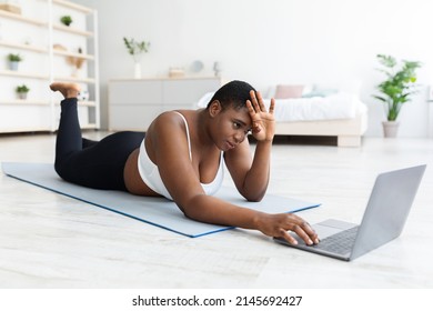 Tired Plus Size Black Woman Lying On Yoga Mat Near Laptop, Wiping Forehead After Strength Training At Home. Millennial Overweight Lady Feeling Exhausted After Online Weight Loss Workout