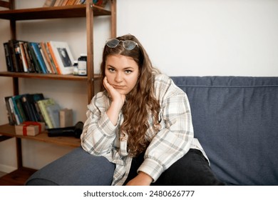 Tired plus size female in casual clothes sitting on couch holding her chin with hand, looking at camera with exhausted facial expression, having rest after finishing household chores - Powered by Shutterstock