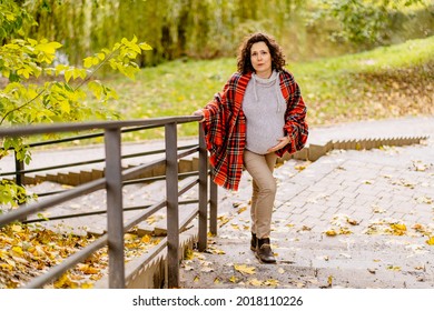 Tired Pregnant Curly Middle Aged Woman In Plaid Scarf And Warm Knitted Sweater With Back Pain Going Up Stairs During Walking In Autumn Park.