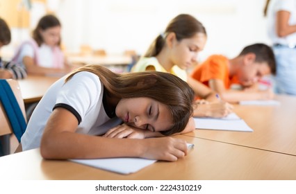 Tired preteen schoolgirl sleeping at desk in classroom during lesson with her head resting on her hands