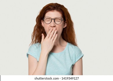 Tired red-headed woman in glasses closed eyes cover mouth with hand yawning feels weak bored pose on grey white background, studio head shot. Chronic fatigue syndrome, lack of sleep and energy concept