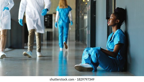 Tired Sad African American Woman Doctor In Uniform And Gloves Taking Off Medical Mask And Leaning On Wall In Despair. Upset Female Nurse Resting, Having Break. Coronavirus. Rest At Work. Covid-19.