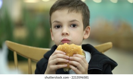 Tired Sad Boy Of Seven Years Old Eats A Burger At A Food Court After A School Day. Child In Sweatshirt Enjoying A Juicy Burger.