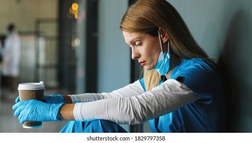 Tired Sad Caucasian Woman Doctor In Medical Mask And Gloves Drinking Coffee And Leaning On Wall In Despair. Upset Female Nurse Resting, Having Break, Sipping Drink. Coronavirus Concept. Rest At Work.