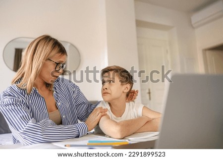 Tired sad kid sits at table with laptop and textbooks while mom help him with homework