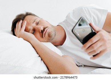 Tired Sad Man With Headache And Illness Symptoms Holding Mobile Phone While Lying In Bed. Frustrated Guy Looking At Screen Of Smartphone While Relaxing In Bedroom Indoors.