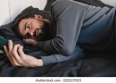 Tired Sad Man With Headache And Illness Symptoms Holding Mobile Phone While Lying In Bed. Frustrated Guy Looking At Screen Of Smartphone While Feel Depression, Or Breaking Up With His Girl, Loneliness