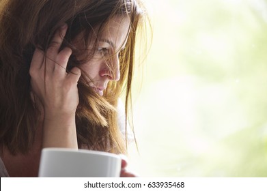 Tired And Sad Woman Is Sitting In The Window Seat With A Cup In Her Hand. Depression Concept. 