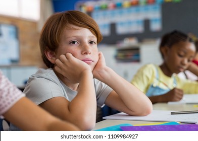 Tired School Boy With Hand On Face Sitting At Desk In Classroom. Bored Schoolchild Sitting At Desk With Classmates In Classroom. Frustrated And Thoughtful Young Child Sitting And Looking Up.