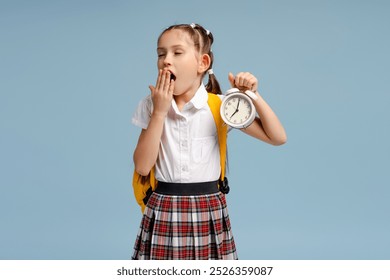 Tired schoolgirl yawning and holding alarm clock while going to school against blue background - Powered by Shutterstock