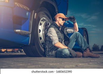 Tired Semi Truck Driver Seating On A Truck Stop Parking Pavement Next To His Tractor Vehicle.