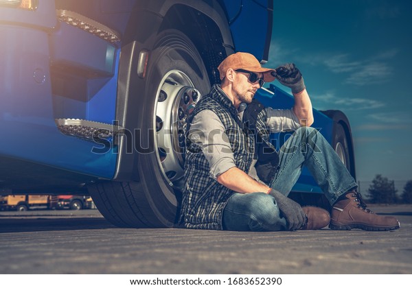 Tired Semi Truck Driver Seating on a\
Truck Stop Parking Pavement Next to His Tractor\
Vehicle.
