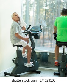 Tired Senior Man With Towel On Exercise Bike In Fitness Club