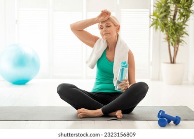 Tired Senior Woman With Water Bottle In Hand Relaxing After Home Sports Training, Athletic Mature Female Resting On Fitness Mat After Workout And Wiping Sweat Off Forehead, Copy Space - Powered by Shutterstock