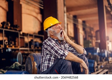 A Tired Senior Worker Is Sitting Next To The Machines And Yawning. After Hard Work, He Must Get Some Sleep. A Sleepy Worker At The Factory.