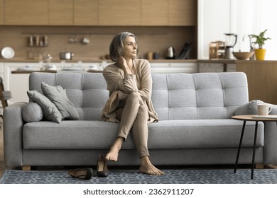 Tired serious middle aged grey haired woman sitting on home couch after long business day, taking shoes off, touching neck, looking away, thinking, feeling fatigue, burnout