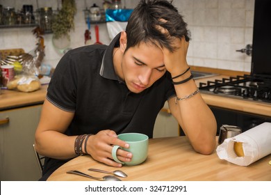 Tired Or Sick Attractive Young Man With Eyes Closed And Hand In Hair, Leaning With Elbow On Kitchen Table And Holding Cup Of Coffee, While Catching Up On Sleep