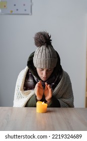 Tired Sick Woman Freezing At Home, Sitting At The Table Warming Hands Of Burning Candle. Woman With Home Heating Problem Feeling Cold. No Heating In Winter At Home.