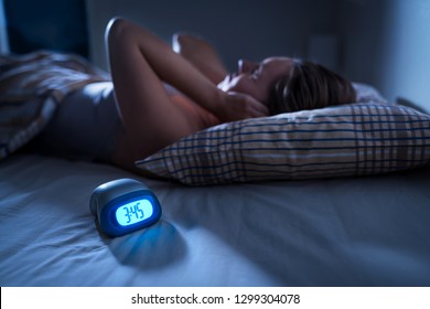 Tired sleepless woman covering ears. Can't sleep. Noisy neighbors, tinnitus, insomnia or stress concept. Noise from party next door. Awake in bed in the middle of the night. Alarm clock with time.  - Shutterstock ID 1299304078