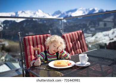 Tired Sleepy Child In Outdoor Restaurant In The Mountains. Family Having Apres Ski Lunch In Alpine Resort. Kids Eat After Skiing. Winter Snow Fun For Child. Food And Drink After Sport. Mountain View.