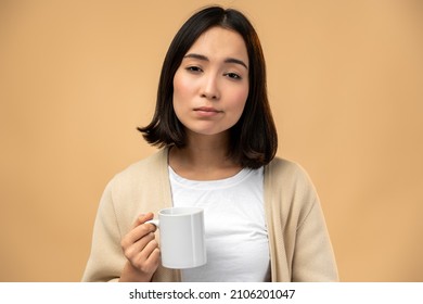 Tired sleepy woman holding cup of coffee, looking at the camera with tired expression, cannot wake up in the morning and go to work. Isolated beige background 
